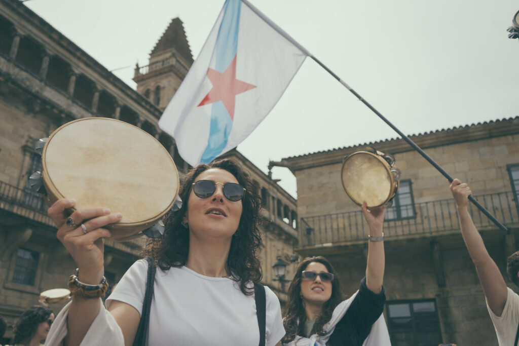 Dúas mulleres erguen pandeiretas. Por detrás vese unha bandeira da patria.