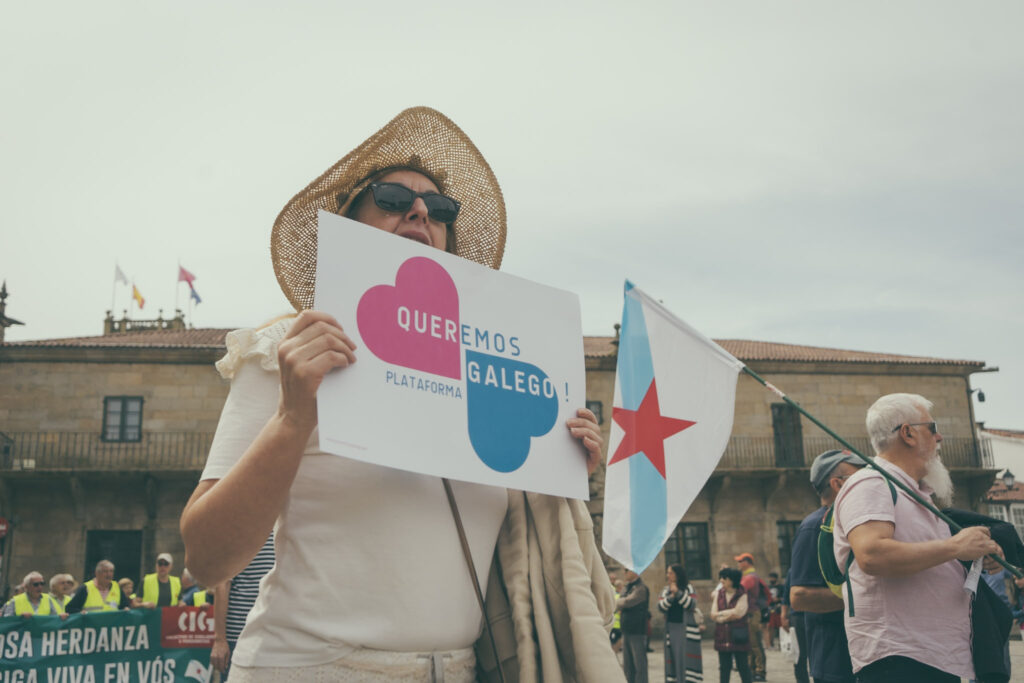 Muller sostén un cartaz co logotipo de Queremos Galego. En segundo plano un home leva unha bandeira da patria.
