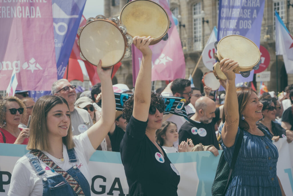 Tres persoas erguen as súas pandeiretas. Ao fondo a multitutde da manifestación.