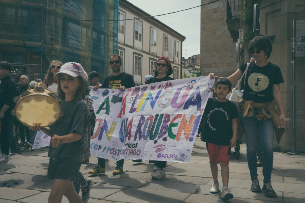 Crianzas e as súas familias sosteñen unha faixa en defensa do galego. Vese tamén algunha crianza tocando a pandeireta.
