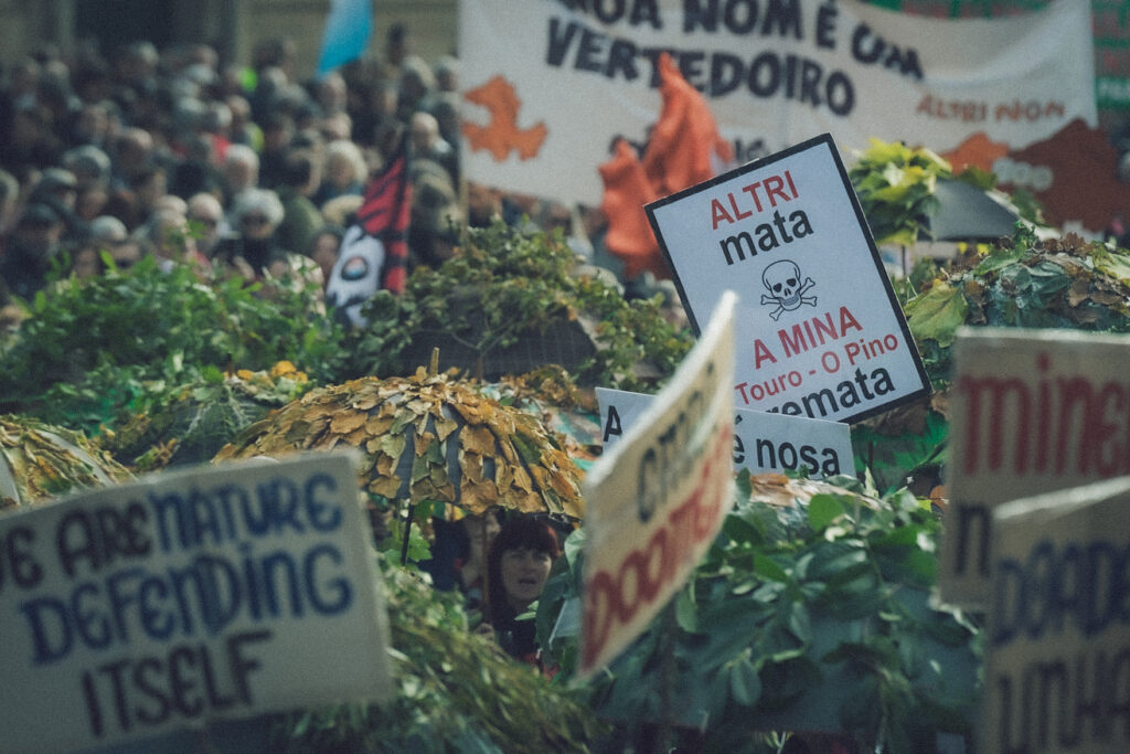 Paraugas con follas de árbores autóctonas. Hai varios cartaces polo medio e destaca un que di "Altri mata, a Mina (Touro - O Pino) remata"