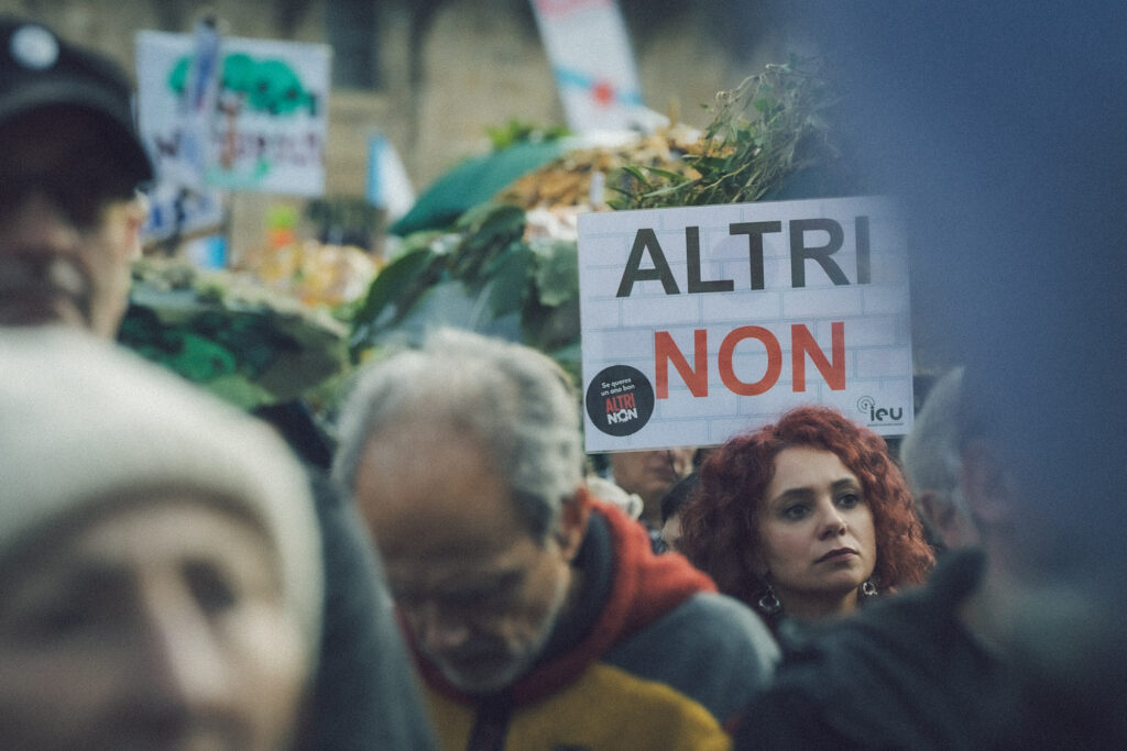 Primeiro plano con caras desenfocadas e ao fondo unha muller enfocada que ten detrás un cartaz que pon ALTRI NON
